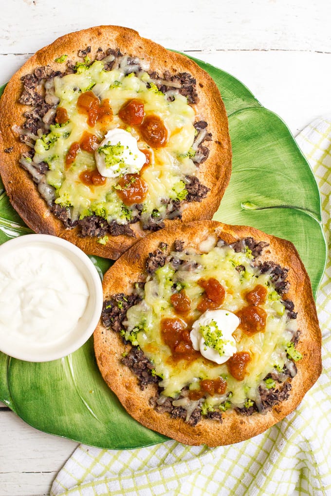 Two homemade baked tostadas topped with mashed black beans, chopped broccoli, melted cheddar cheese and salsa and sour cream on top.