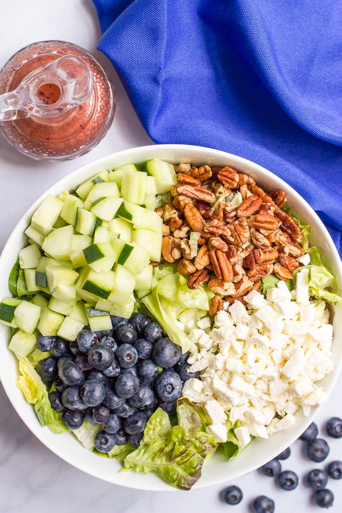A salad being assembled in a bowl full of lettuce with diced cucumber, chopped pecans, crumbled feta and fresh blueberries on top.