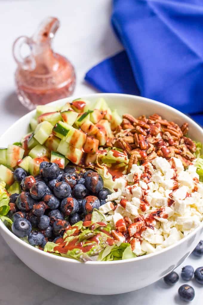 A white bowl with a salad with separate piles of cucumbers, blueberries, pecans and feta cheese and a blueberry vinaigrette drizzled on top.