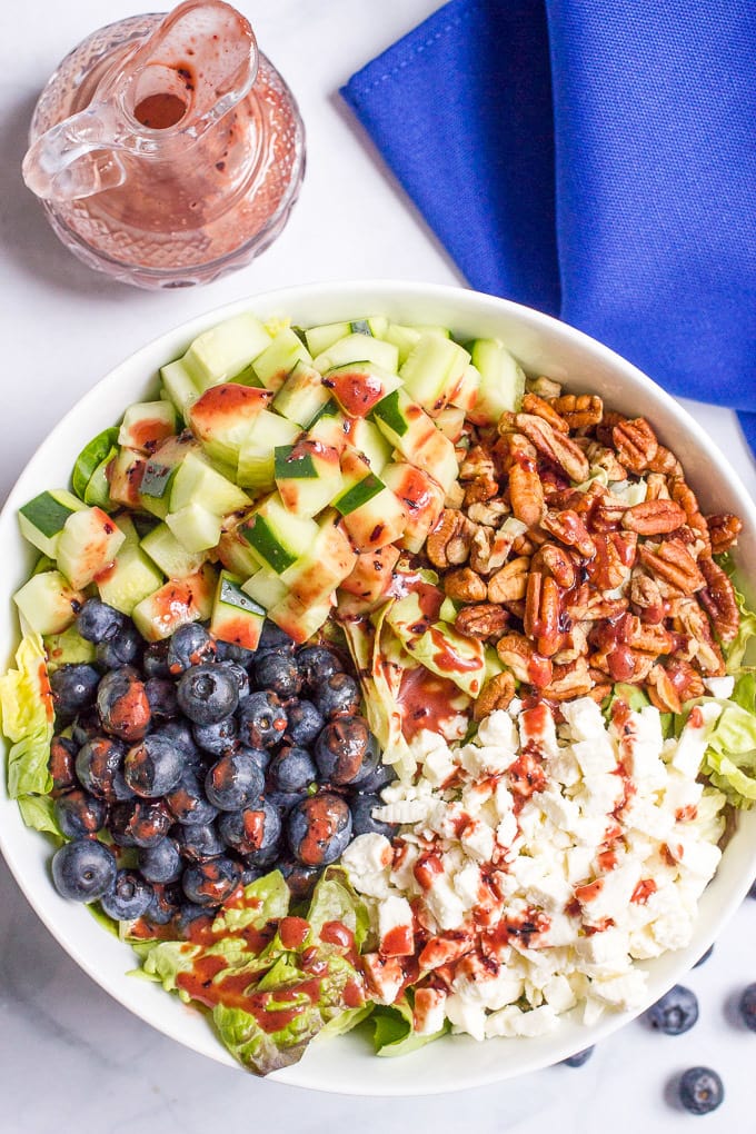 A white bowl with a salad with separated piles of diced cucumber, blueberries, pecans and feta cheese and a blueberry vinaigrette drizzled on top.