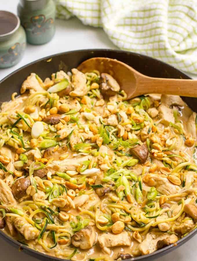 A wooden spoon resting in a large skillet with chicken, mushrooms and zucchini noodles in a satay sauce.