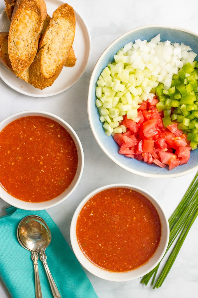 Bowls of gazpacho with extra chopped veggies and toasted bread for garnishes in separate bowls nearby