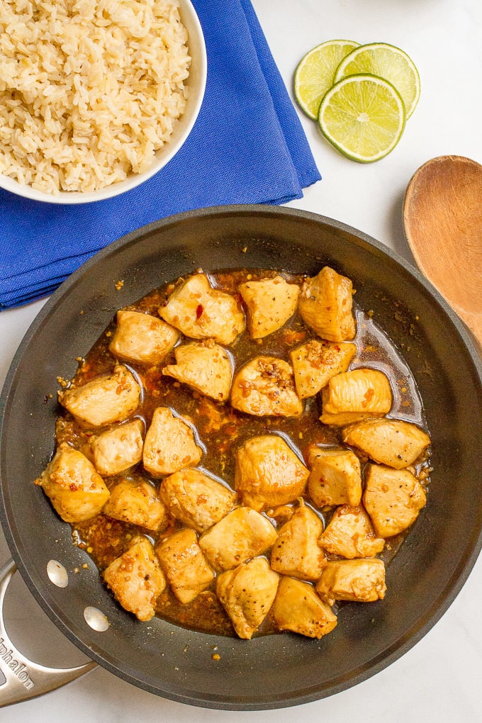 Chicken cubes in a honey garlic sauce in a dark skillet with a wooden spoon to the side, lime wedges nearby and a bowl of rice for serving