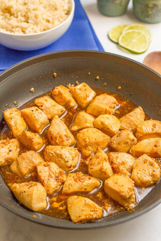 A dark skillet with cooked cubed chicken pieces in a sauce and a bowl of rice in the background