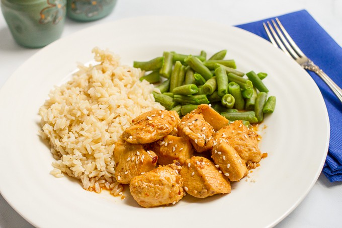 Honey garlic chicken topped with sesame seeds and served with rice and green beans on a white round dinner plate