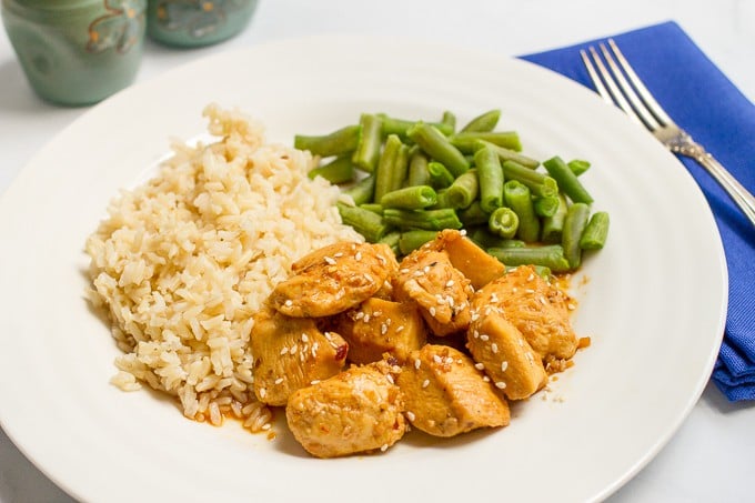 Honey garlic chicken topped with sesame seeds and served with rice and green beans on a white round dinner plate