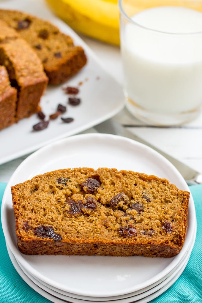 A slice of homemade cinnamon raisin bread on a small white plate with the rest of the bread and a glass of milk in the background.