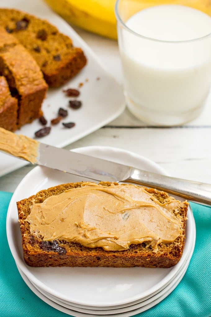 A slice of homemade cinnamon raisin bread on a small white plate with peanut butter smeared over the top and the rest of the bread and a glass of milk in the background.