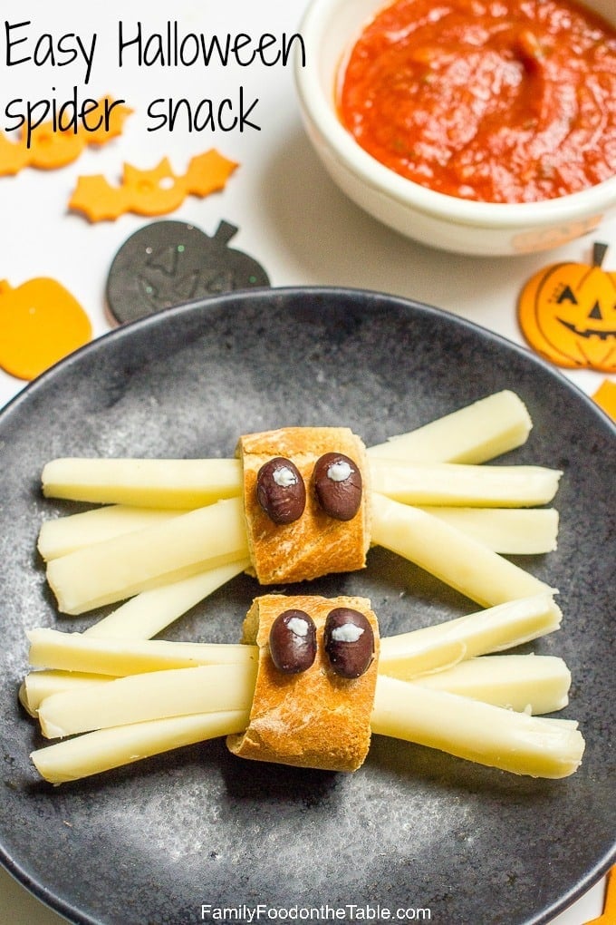 Close up of string cheese spider snacks on a black plate with Halloween themed sticker scattered about and a bowl of marinara for dipping in the background.