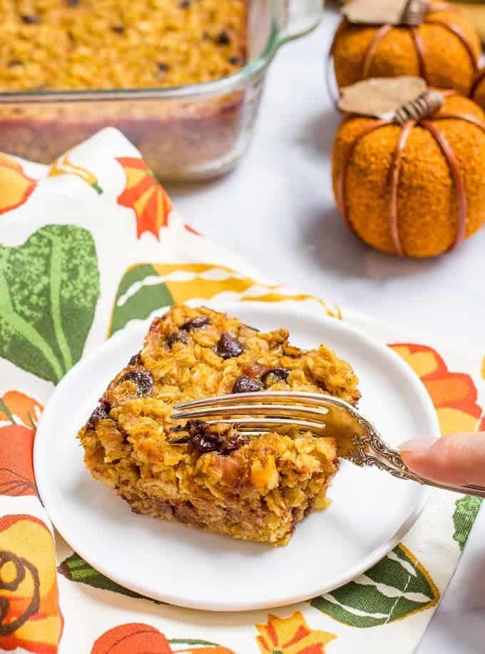 A fork cutting into a piece of baked oatmeal with pumpkin and chocolate chips on a small white plate.