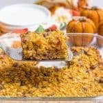 A spatula lifting up a slice of pumpkin baked oatmeal with chocolate chips from a glass baking dish with faux pumpkins and a stack of plates in the background.