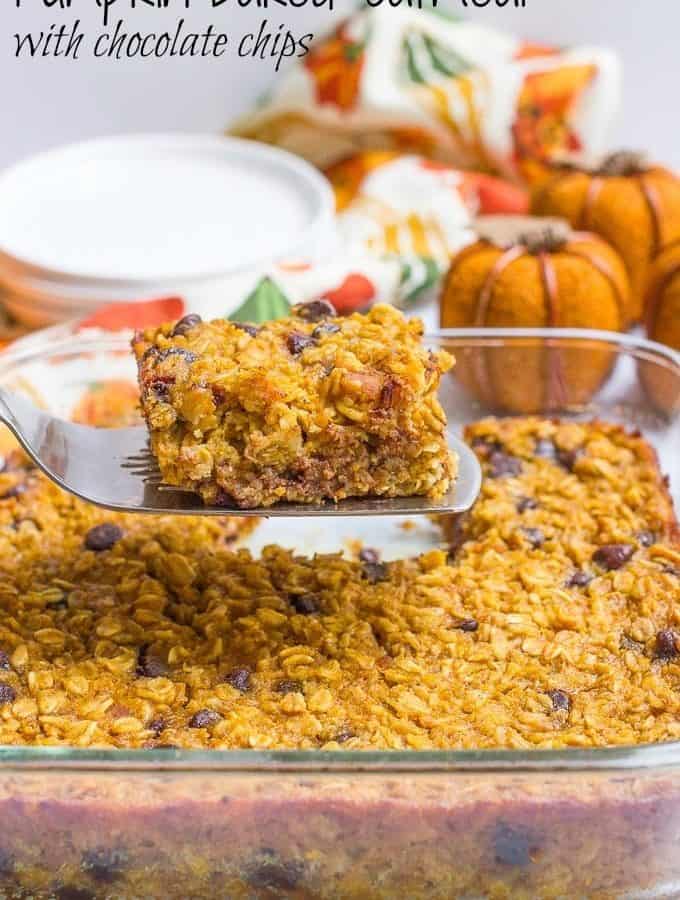 A spatula lifting up a slice of pumpkin baked oatmeal with chocolate chips from a glass baking dish with faux pumpkins and a stack of plates in the background.