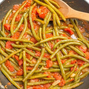 A wooden spoon resting in a skillet with a mix of sautéed green beans with canned tomatoes.