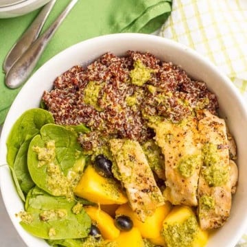 A white bowl with separate piles of red quinoa, sliced chicken, mango and blueberries and baby spinach with a dressing drizzled over top.