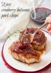 A hand spoon some cranberry balsamic sauce over pork chops on a plate with a glass of red wine in the background.