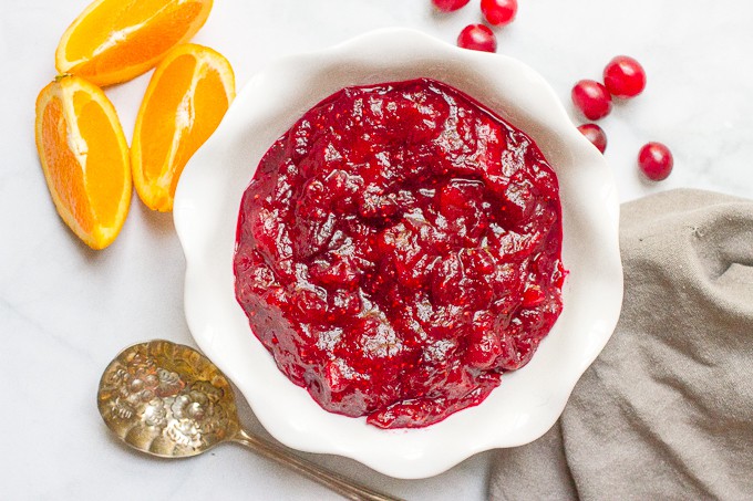 A fluted white bowl with chunky homemade cranberry sauce, with orange slices and fresh cranberries nearby