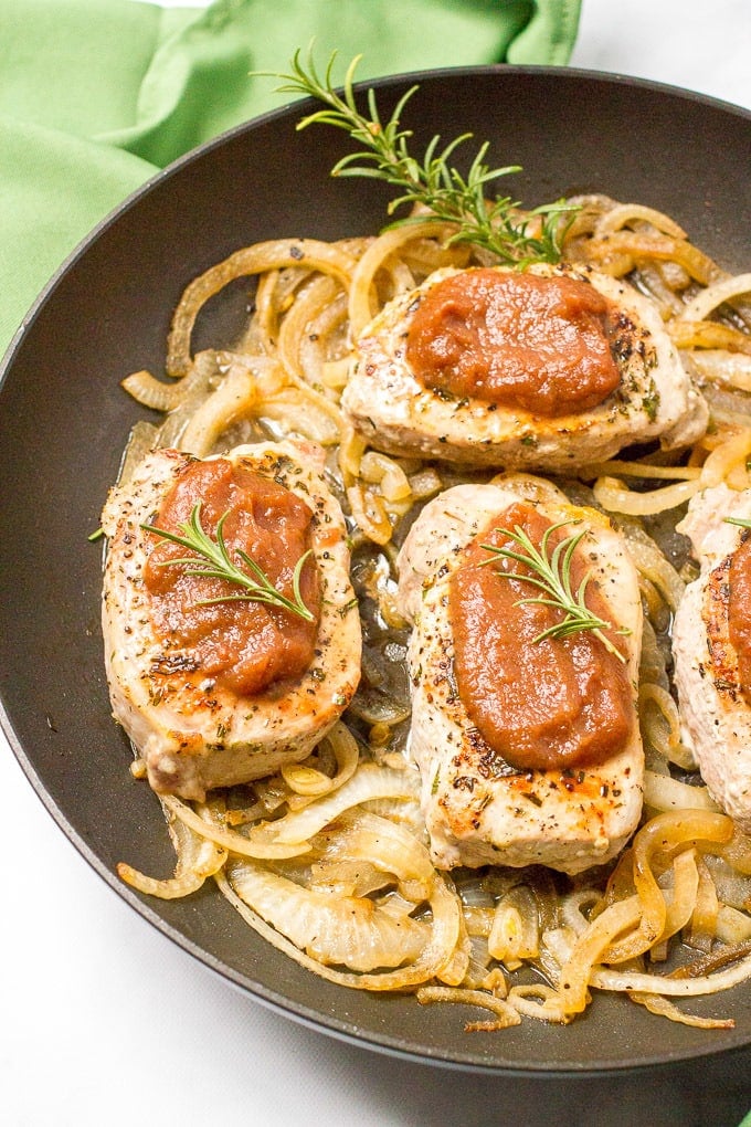 A large skillet with sautéed onions and seared pork chops topped with apple butter and sprigs of rosemary.