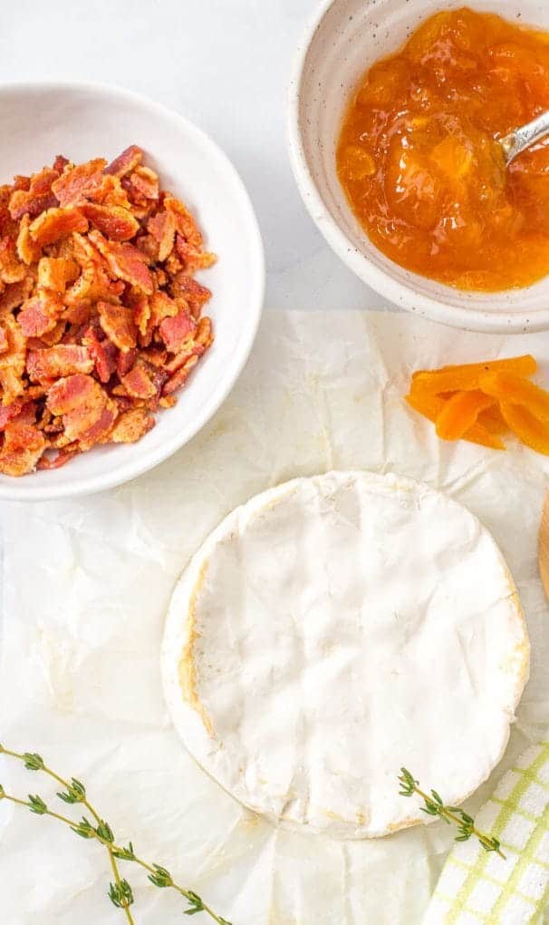 A wheel of brie cheese alongside apricot jam, slivered apricots and a bowl of cooked, crumbled bacon.