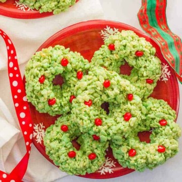 A red holiday plate with a stack of green dyed wreath shaped rice krispie treats dotted with red hots for decoration.