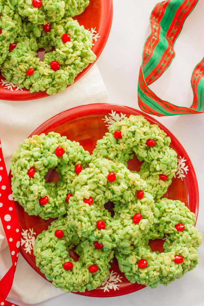 A red holiday plate with a stack of green dyed wreath shaped rice krispie treats dotted with red hots for decoration with holiday wrapping ribbons nearby.