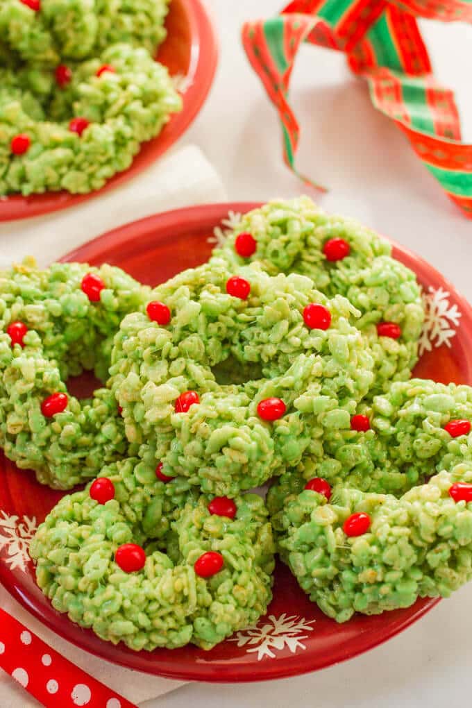 Rice Krispie treats dyed green and shaped like wreaths and decorated with red hots candies on a red plate.
