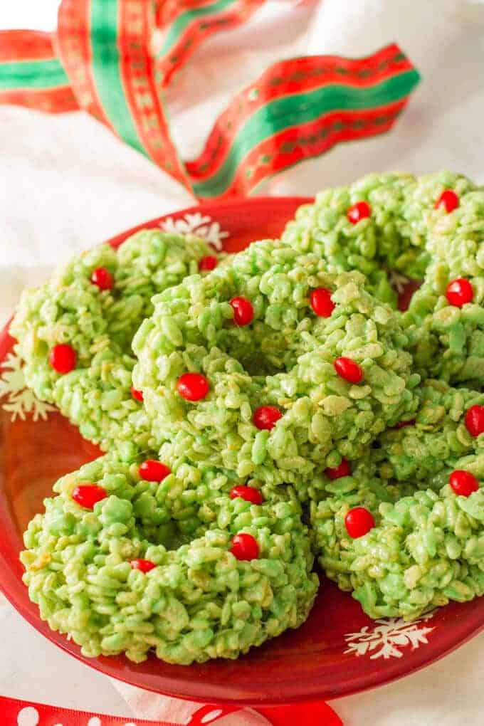 Close up of a stack of green dyed wreath shaped rice krispie treats dotted with red hots for decoration on a red holiday themed plate.
