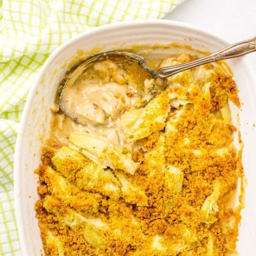 A large serving spoon resting in a white baking dish with a creamy turkey artichoke casserole topped with browned breadcrumbs.