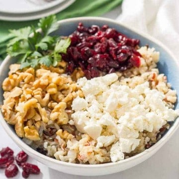 A blue tinted bowl full of a wild rice mixture topped with pecans, dried cranberries and feta cheese before being mixed in with extra dried cranberries to the side.