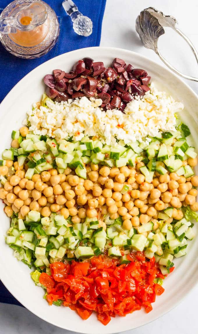 Rows of salad ingredients in a bowl before being mixed together, including olives, feta cheese, cucumber, chickpeas and roasted peppers.