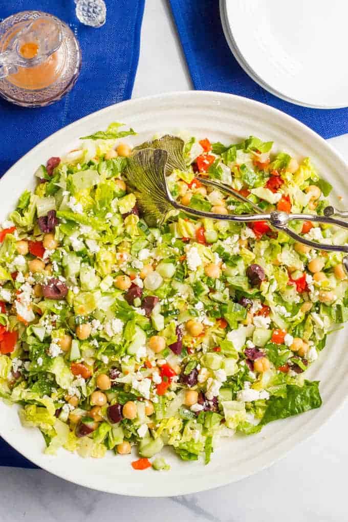 A Mediterranean chopped salad with lettuce, cucumber, olives, peppers, chickpeas and feta cheese in a large white serving bowl with silver tongs resting in it.