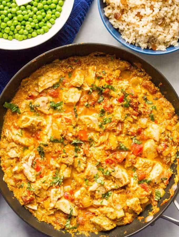 A skillet with a saucy chicken curry and bowls of rice and peas behind it