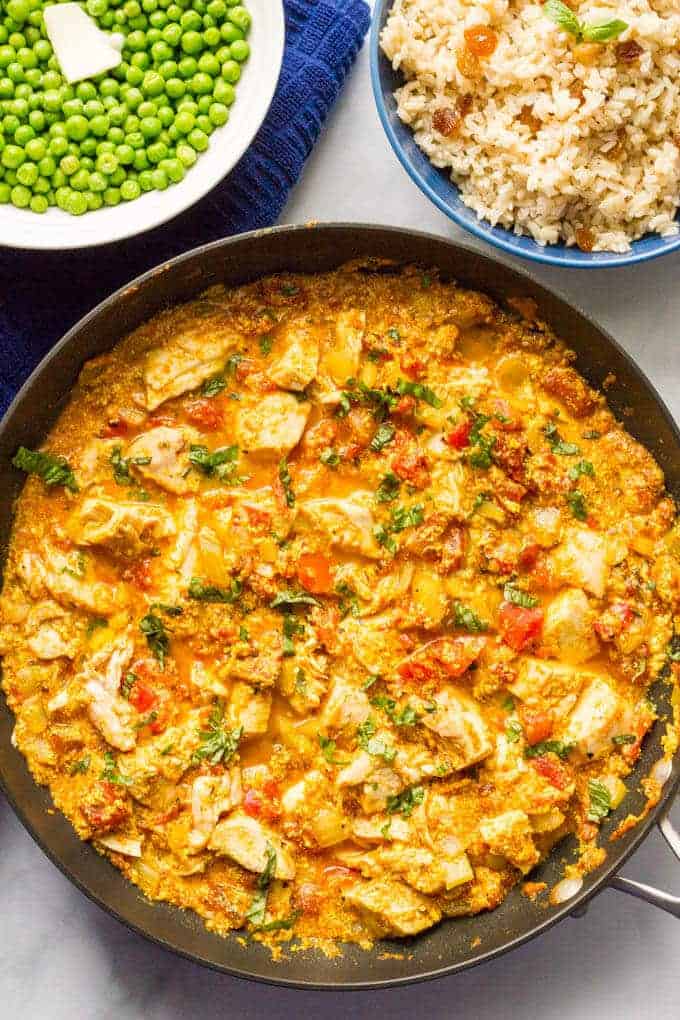 A skillet with a saucy chicken curry and bowls of rice and peas behind it