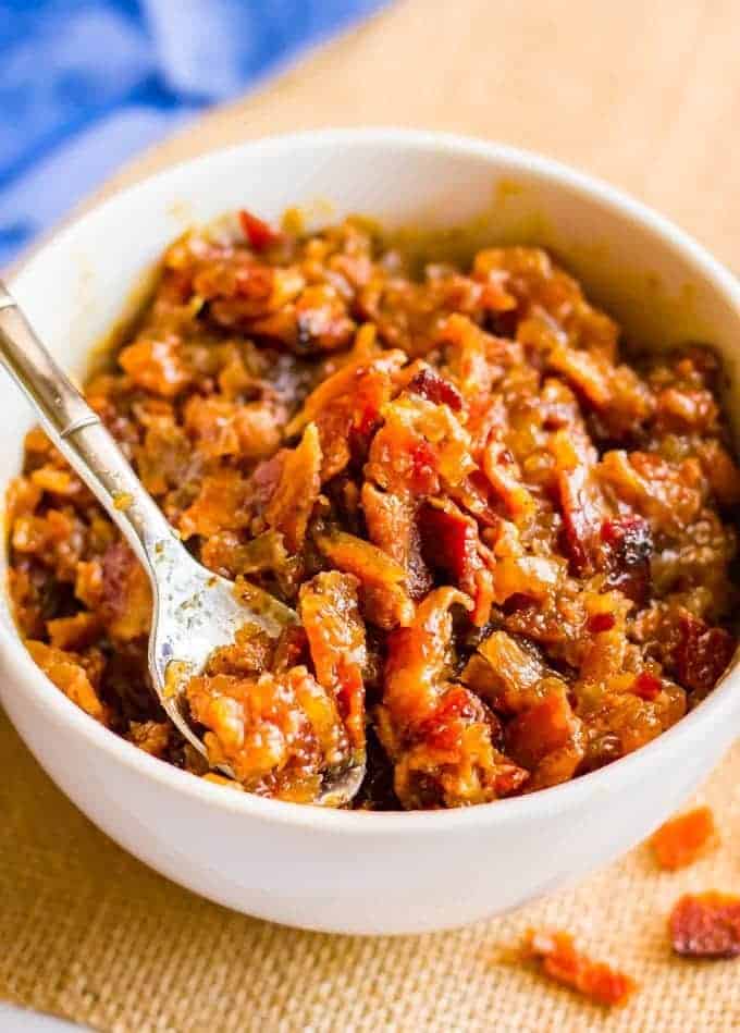 A spoon resting in a bowl of bourbon bacon jam with extra bacon pieces nearby