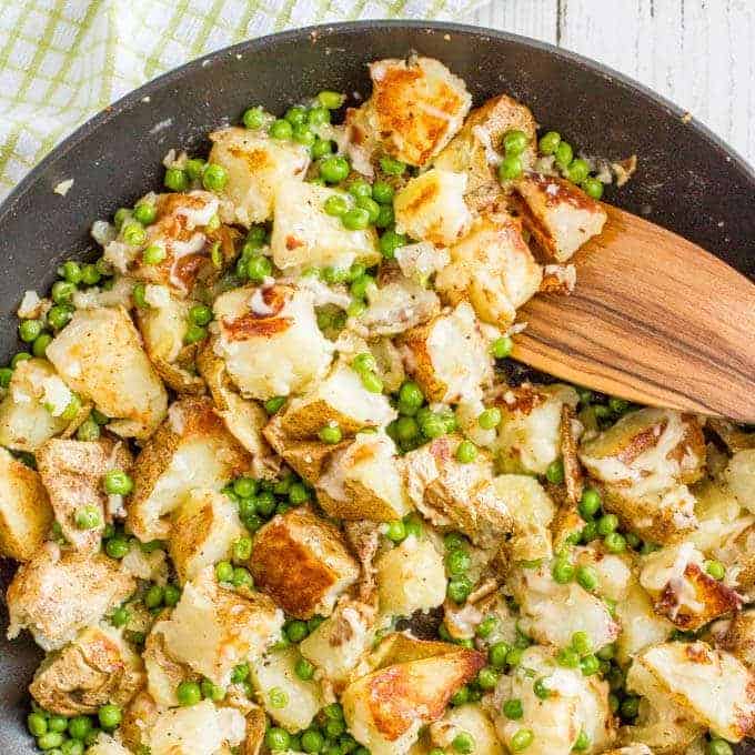 A wooden spoon stirring a skillet full of cheesy potatoes and peas