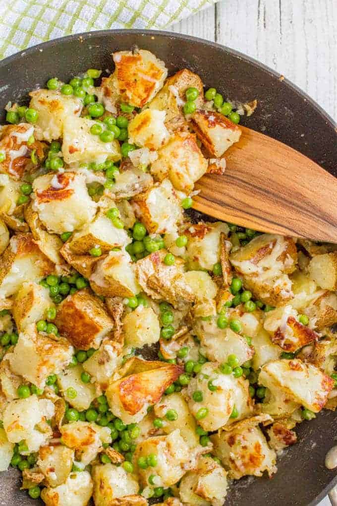 A large skillet with browned potatoes and green peas and a wooden spoon on the side