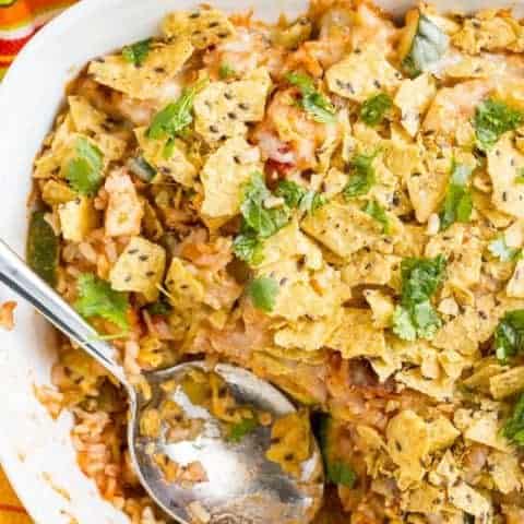 A white casserole dish with a chicken taco mixture topped with cheese and tortilla chips and a spoon resting in the dish