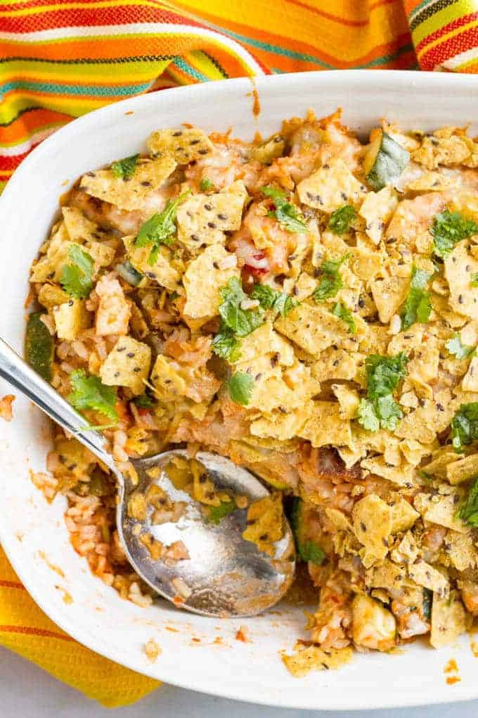 A white casserole dish with a chicken taco mixture topped with cheese and tortilla chips and a spoon resting in the dish