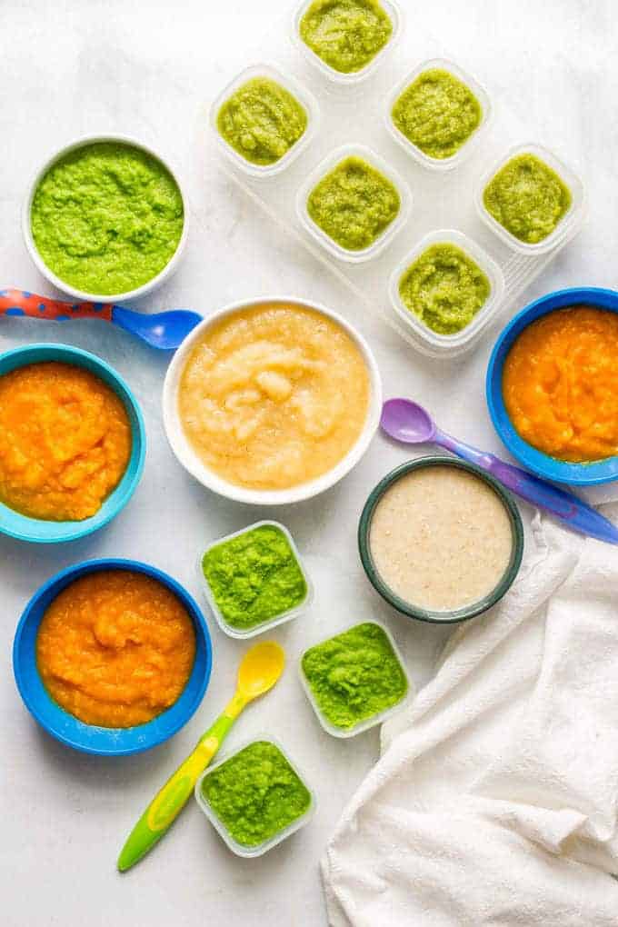 A big variety of different colorful homemade baby foods in a variety of different sized bowls and containers with baby spoons scattered around.