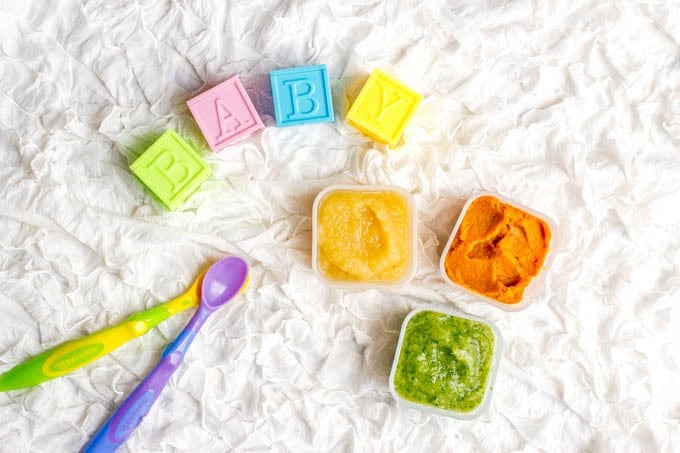 A trio of small containers of homemade baby food on a ruffled white blanket background with baby spoons and baby blocks nearby.