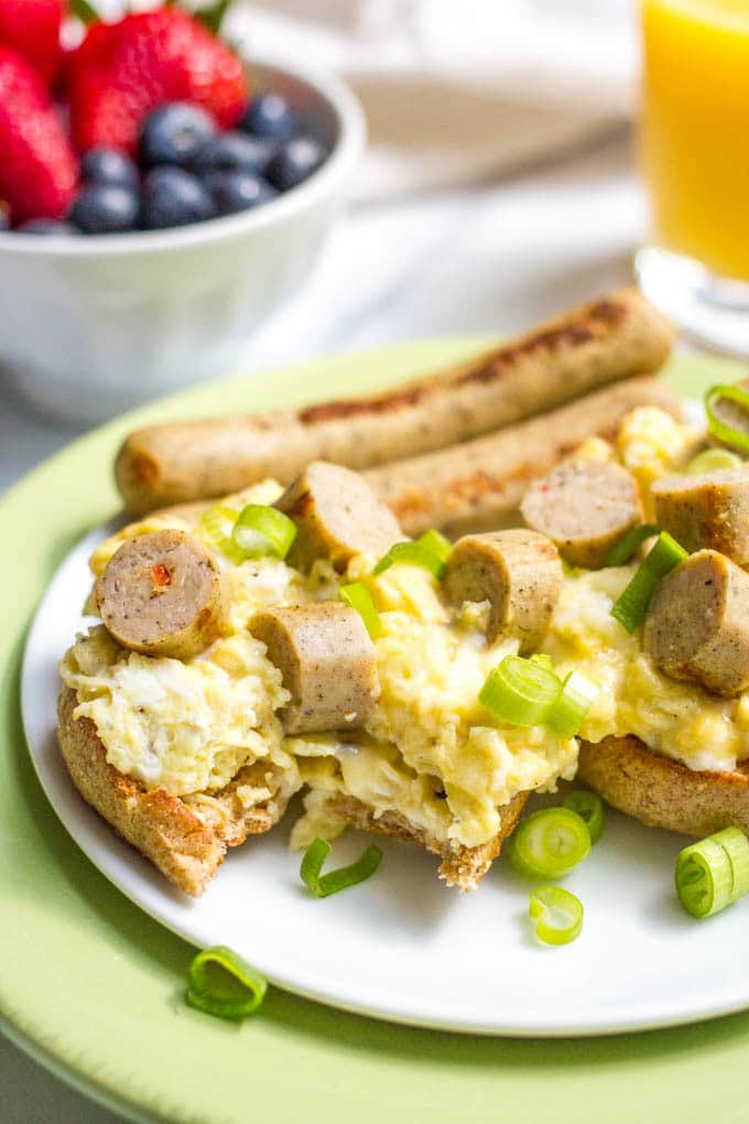 A halved bagel with a bite taken out that's topped with cheesy scrambled eggs and slices of chicken breakfast sausages with green onions