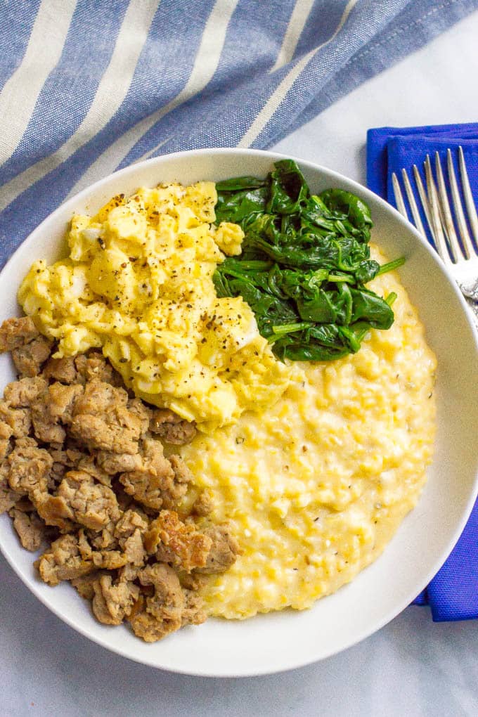 A white bowl filled with cheesy grits topped with crumbled sausage, scrambled eggs and sautéed spinach