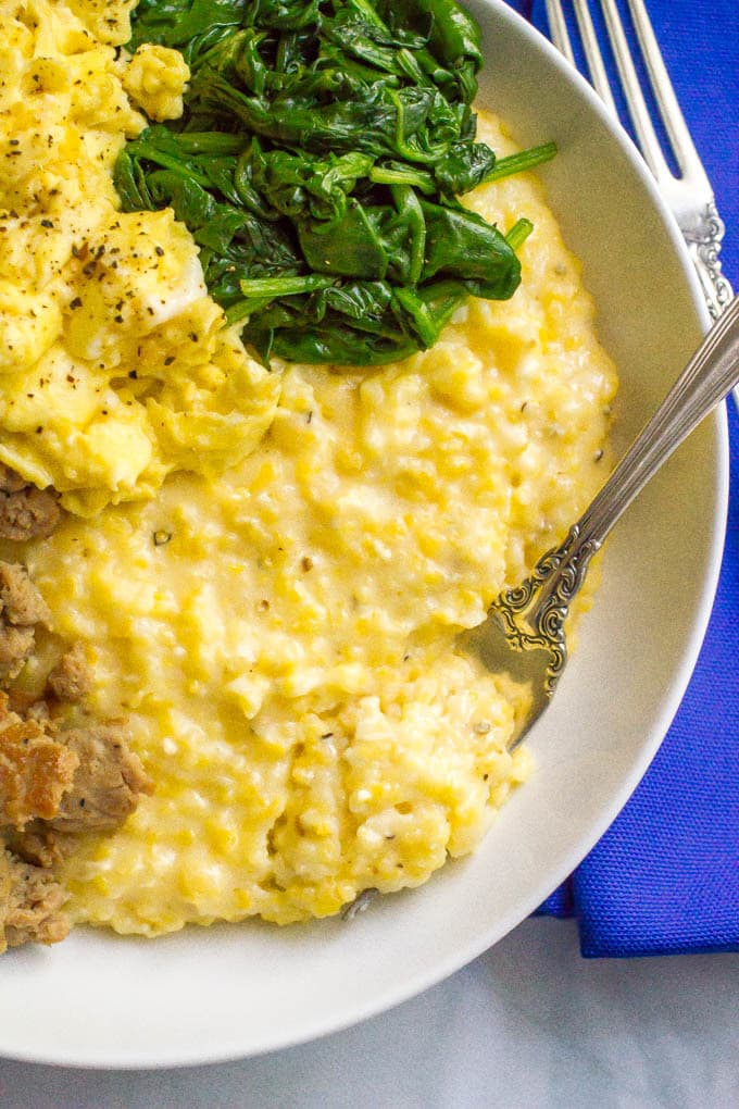 Close up of a fork resting in a bowl of cheesy grits with sausage, eggs and spinach to the side of the bowl