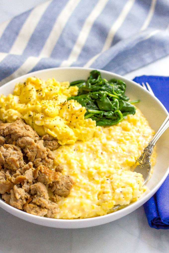 A fork resting in a bowl of cheesy grits with crumbled sausage, scrambled eggs and sautéed spinach