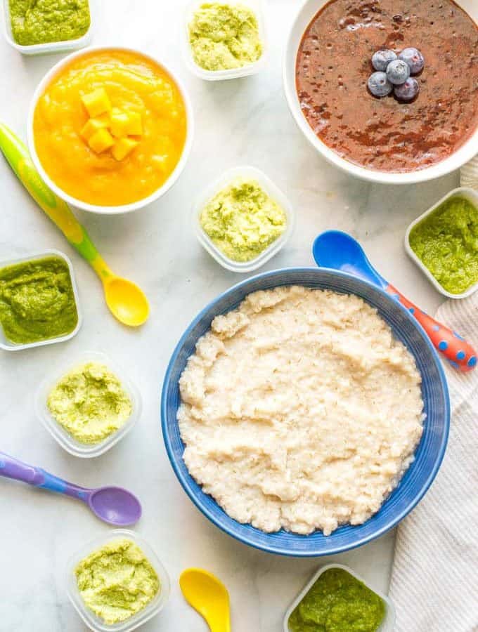 Separate bowls and small containers of different homemade baby food purees, including apricot and blueberry purees, with colorful baby spoons scattered about.