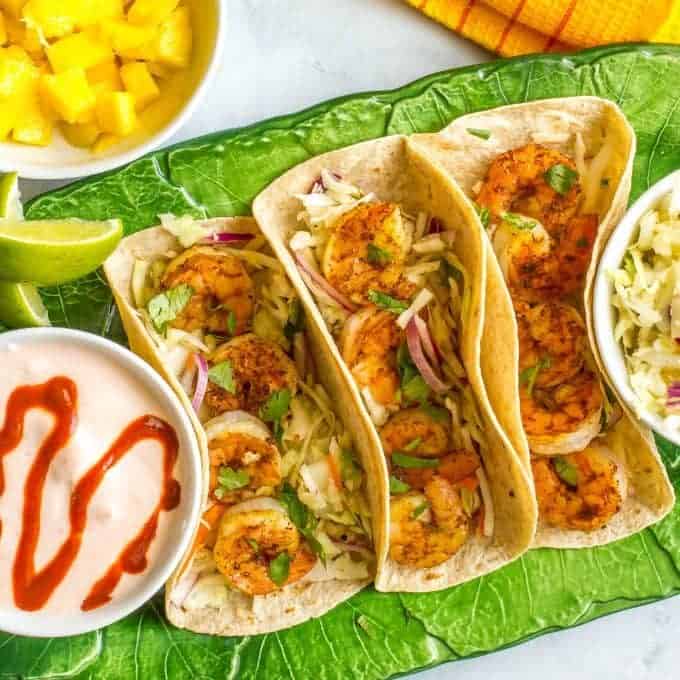 Three shrimp tacos in flour tortillas set on a green serving platter with bowls of sauce and a cabbage slaw to the side with fresh pineapple in a bowl in the background.