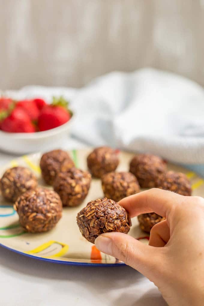 A hand picking up a chocolate cookie ball from a plate and a bowl of strawberries in the background.