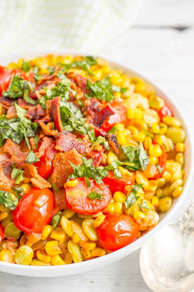 A large white serving bowl of succotash with bacon and fresh herbs