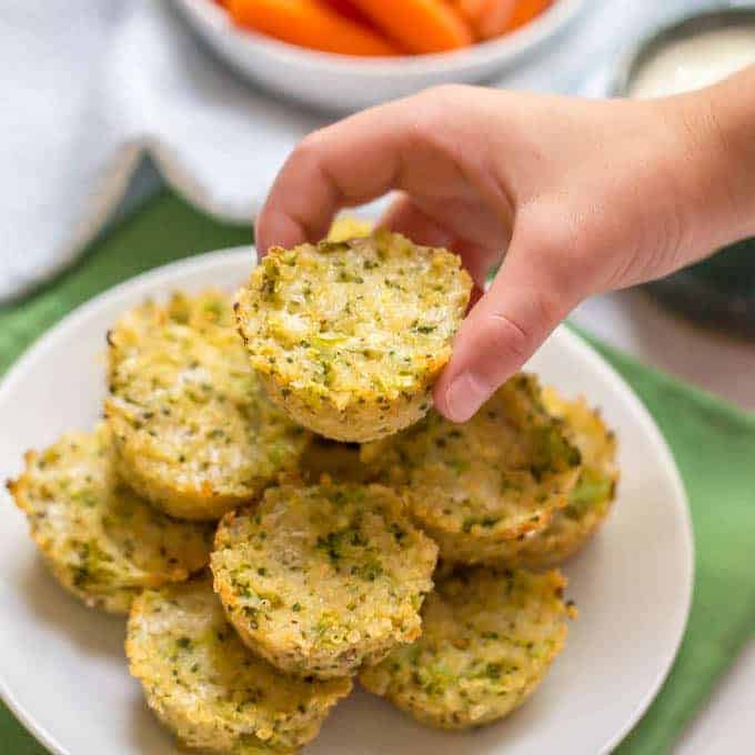 A hand picking up a cheesy broccoli mini muffin from a stack on a plate.