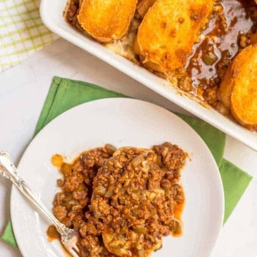 A small white plate with a serving of cheesy sloppy Joe casserole and the entire casserole dish beside it