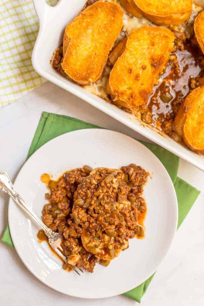 A small white plate with a serving of cheesy sloppy Joe casserole and the entire casserole dish beside it